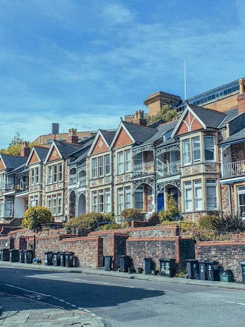 A row of Victorian-style terraced houses on a residential street in Hammersmith, featuring ornate white decorative trim and bay windows. The houses are constructed with red brick and have pitched roofs with dark tiles. Small front gardens with neatly trimmed bushes and greenery are situated behind low brick walls. The street is paved with asphalt, with black wheelie bins lined up along the sidewalk. Bright daylight and a clear blue sky illuminate the scene. The image emphasizes residential property maintenance and cleanliness, aligning with deep surface cleaning and sanitisation themes discussed on the page for avoiding hidden cleaning fees, as provided by Carpet Cleaning Hammersmith.