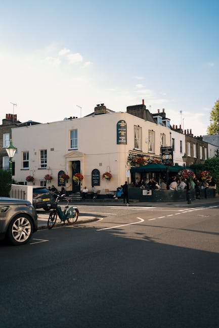 A street scene featuring a white, multi-story building with large windows and a dark green awning, located at a busy corner in Hammersmith. The building's exterior is decorated with hanging flower baskets, and outdoor seating is visible, suggesting a café or restaurant. The pavement and road are clean, with a black car parked in the foreground and a bicycle leaned against a lamppost. Bright daylight and clear sky lighting highlight the polished surfaces and well-maintained appearance of the surroundings. As part of professional cleaning services, Carpet Cleaning Hammersmith emphasizes the importance of surface cleaning and deep cleaning to maintain hygiene and cleanliness in such commercial and residential settings, ensuring surfaces like windows, facades, and outdoor furniture are well sanitised and free from dust and grime.