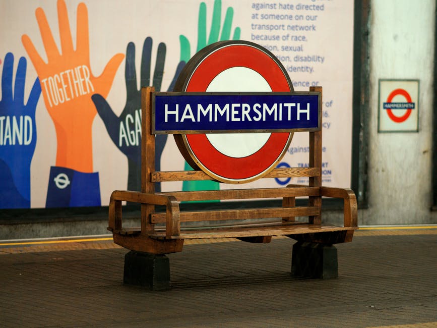 A wooden bench with a slatted seat and armrests, positioned on a tiled station platform, features a blue and white sign reading 'HAMMERSMITH' attached to a large round London Underground red and white roundel. In the background, a colorful mural displays multiple hands in orange, green, and dark blue, with the words 'Togeth...'. The scene is lit with soft, natural interior lighting, and the surroundings appear clean and well-maintained, emphasizing the importance of hygiene and surface cleaning in public transport environments, consistent with carpet cleaning and sanitisation standards discussed on the page titled 'Carpet disposal rules in Hammersmith and Fulham council, Hammersmith' at Carpet Cleaning Hammersmith.
