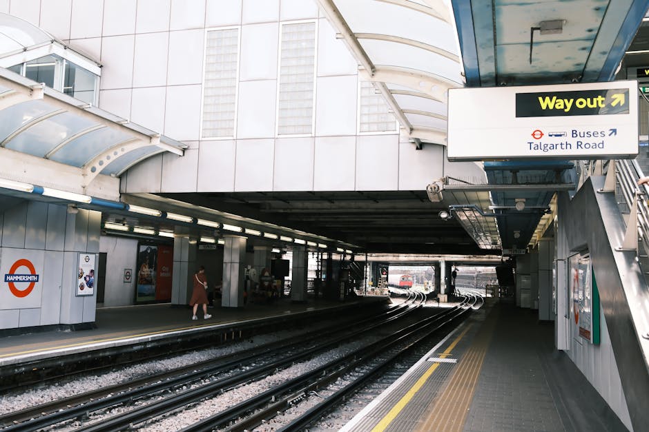 An underground station platform at Hammersmith with silver and white tiled walls, illuminated by fluorescent lighting. The platform features train tracks at the center, with a lowered ceiling and an overhead sign indicating the way out and bus connections at Talgarth Road. Two passengers are visible, one walking and another standing near the edge. There are various posters and signage on the walls, including a prominent Hammersmith station logo, contributing to a clean and well-maintained environment consistent with surface cleaning and hygiene standards. The scene is well-lit, showing an orderly, dust-free space prepared for daily use, representative of the importance of surface cleaning in public transportation areas, as highlighted on the Carpet Disposal Rules in Hammersmith and Fulham council page by Carpet Cleaning Hammersmith.