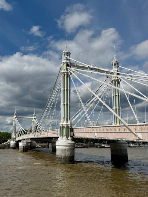 The image depicts a large suspension bridge spanning a river, with tall, ornate towers connected by multiple steel cables and a pale pink decking. The bridge is constructed from metal and stone bases, with the river water visible beneath the structure. The sky above is partly cloudy, with sunlight illuminating parts of the bridge and surrounding area, highlighting the clean, well-maintained appearance of the surfaces. The scene is set during daylight hours, emphasizing the bright atmosphere and the structural details of the bridge. This setting aligns with themes of surface cleaning and maintenance, similar to the deep cleaning and sanitisation tips discussed for mattress cleaning and mould removal at [PAGE_TITLE], offered by [COMPANY_NAME].