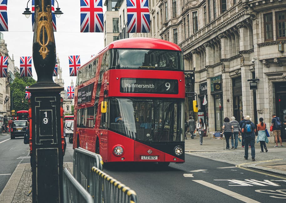 A red double-decker bus displaying route number 9 and destination Hammersmith travels along a city street lined with historic stone buildings and shopfronts. The street is decorated with multiple Union Jack flags hanging from posts, creating a festive atmosphere. Pedestrians, some carrying shopping bags and others wearing backpacks, walk on the sidewalk. In the foreground, a black lamppost with gold decorative elements stands beside a metal barrier with yellow and black striped markings. The scene is illuminated by natural daylight, highlighting the urban environment typical of Hammersmith, London. This image represents a busy city scene with public transport and pedestrian activity.