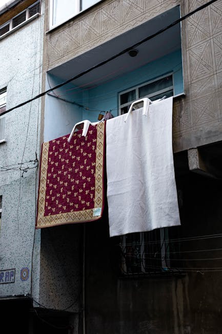 Exterior view of a residential building in Hammersmith with a concrete facade and textured tile detailing on the upper level. A clothesline stretches across the image, suspending a red and gold patterned rug and a plain white towel, both hanging to dry. The window on the upper floor features sliding glass panes with metal frames, partially open, under a light blue awning. The building's surface shows signs of weathering, and the lighting suggests an overcast day. The image highlights urban household laundry hanging in a typical residential setting, emphasizing outdoor maintenance in a city environment. Carpet Cleaning Hammersmith offers professional cleaning and surface sanitisation services for homes and businesses, ensuring hygiene and cleanliness in everyday spaces.