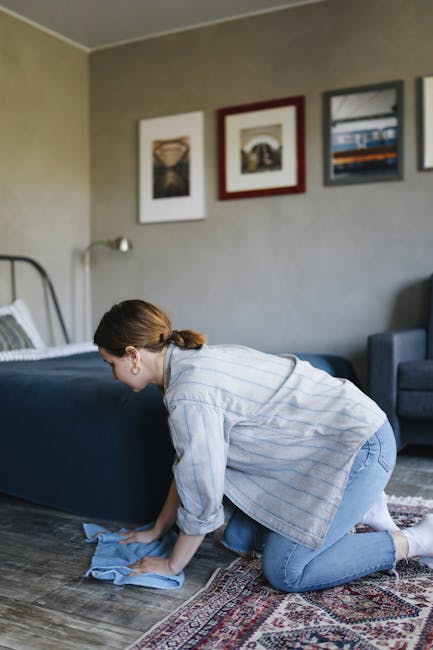 A woman with brown hair tied back, dressed in a light blue striped shirt and jeans, is kneeling on a patterned area rug while using a blue cloth to carefully clean the wooden floor of a bedroom. The room features a bed with a grey metal frame, neatly made with white bedding, and is adorned with three framed artworks arranged horizontally on a beige wall. To the right, there is a dark blue armchair, and above the bed, a small metallic reading lamp is attached to the wall. Natural light appears to illuminate the space, emphasizing a sense of cleanliness and hygiene. This scene showcases domestic surface cleaning and deep cleaning activities, highlighting the professional cleaning services provided by Carpet Cleaning Hammersmith for maintaining a hygienic and well-maintained living environment in W6, London.