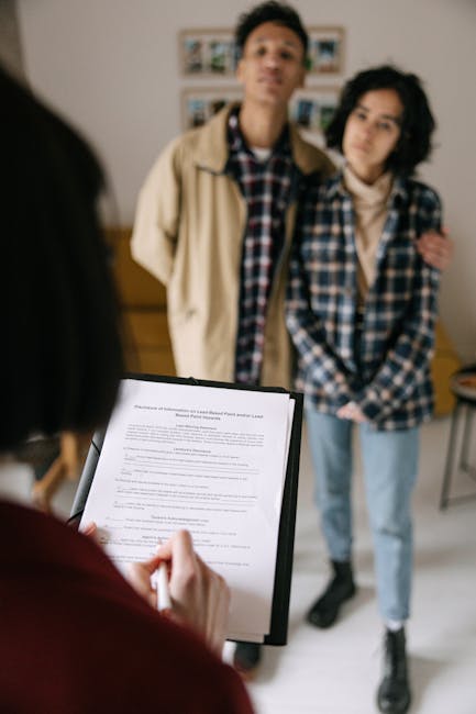 A blurred indoor scene showing a young man and woman standing together in a room with natural light, while a person in the foreground holds a clipboard with documents. The room features neutral-colored walls, and behind the standing individuals, there is a wooden sideboard and a framed photo gallery. The focus is on the people, with no visible surfaces or cleaning tools present, emphasizing a social or testimonial setting related to cleaning services or tenancy discussions, as mentioned on the page about end of tenancy cleaning checklists for landlords and agents. Carpet Cleaning Hammersmith offers expert domestic cleaning and surface sanitisation in such environments.