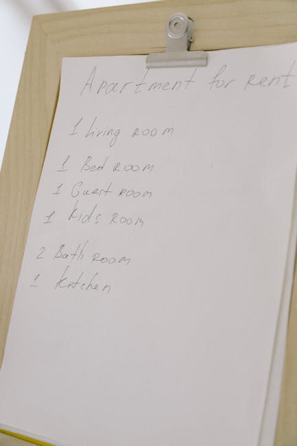 A white paper listing an end of tenancy cleaning checklist for apartments, pinned to a light wood door with a metal clip. The checklist includes rooms such as the living room, bedroom, guest room, kids' room, bathrooms, and kitchen, handwritten in light pencil. The door's surface appears smooth and clean, with no visible dust or damage. The lighting is soft and even, highlighting the neat handwritten notes. This image is relevant to surface cleaning and maintenance tasks for landlords and agents, fitting within the context of domestic cleaning services provided by Carpet Cleaning Hammersmith, as detailed on their website page about the end of tenancy cleaning checklist for Hammersmith.